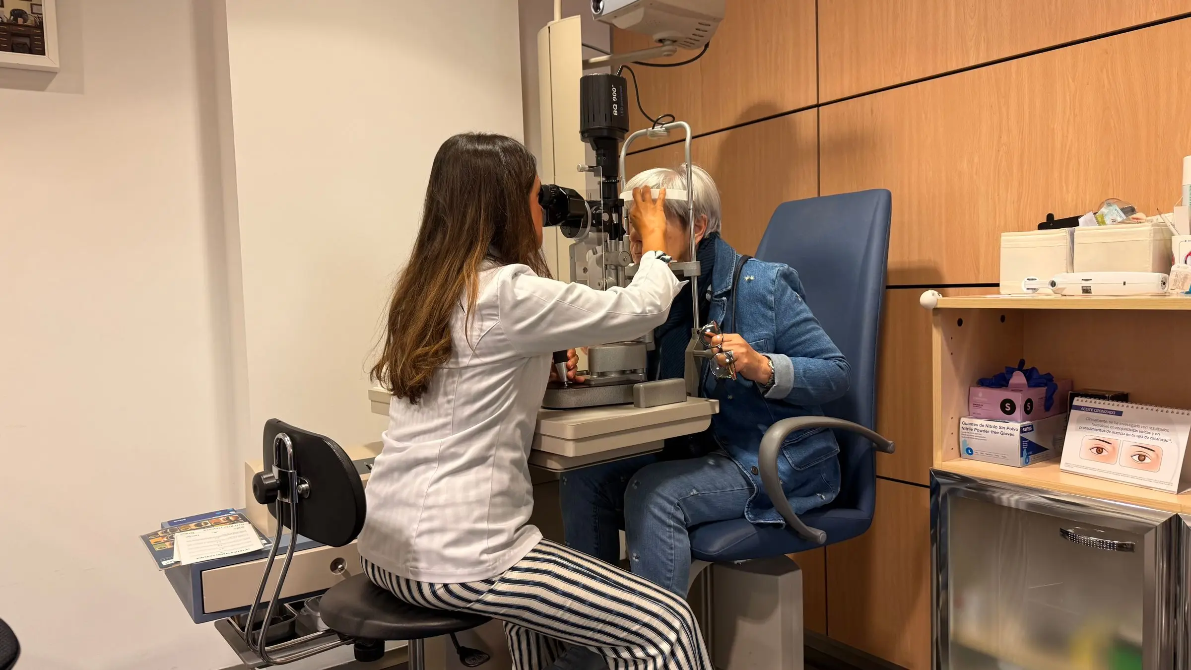 Una optometrista explora con la lámpara de hendidura a una paciente mayor sentada en el sillón del gabinete